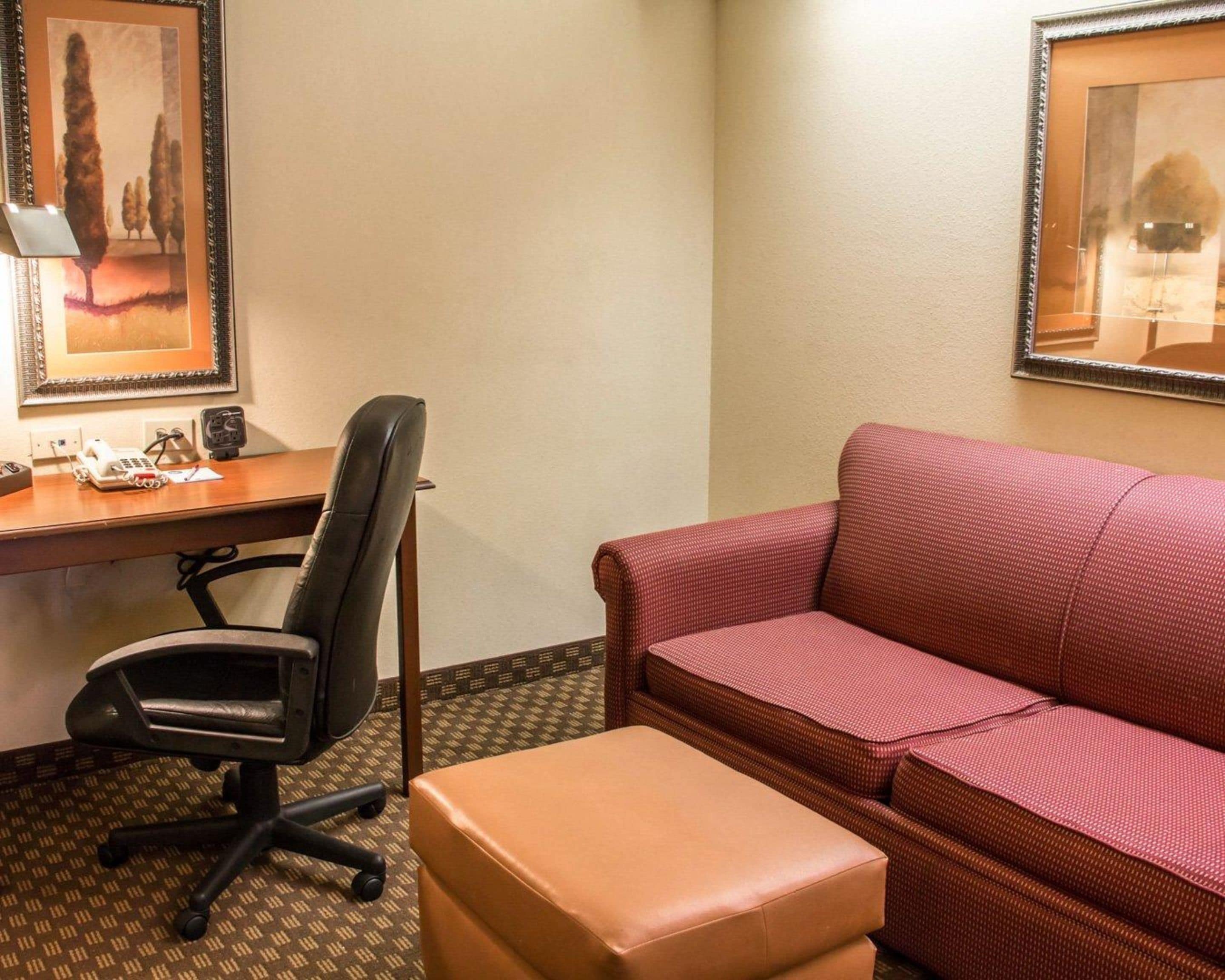 A hotel room with a desk, a black office chair, a red patterned sofa, and a brown ottoman.