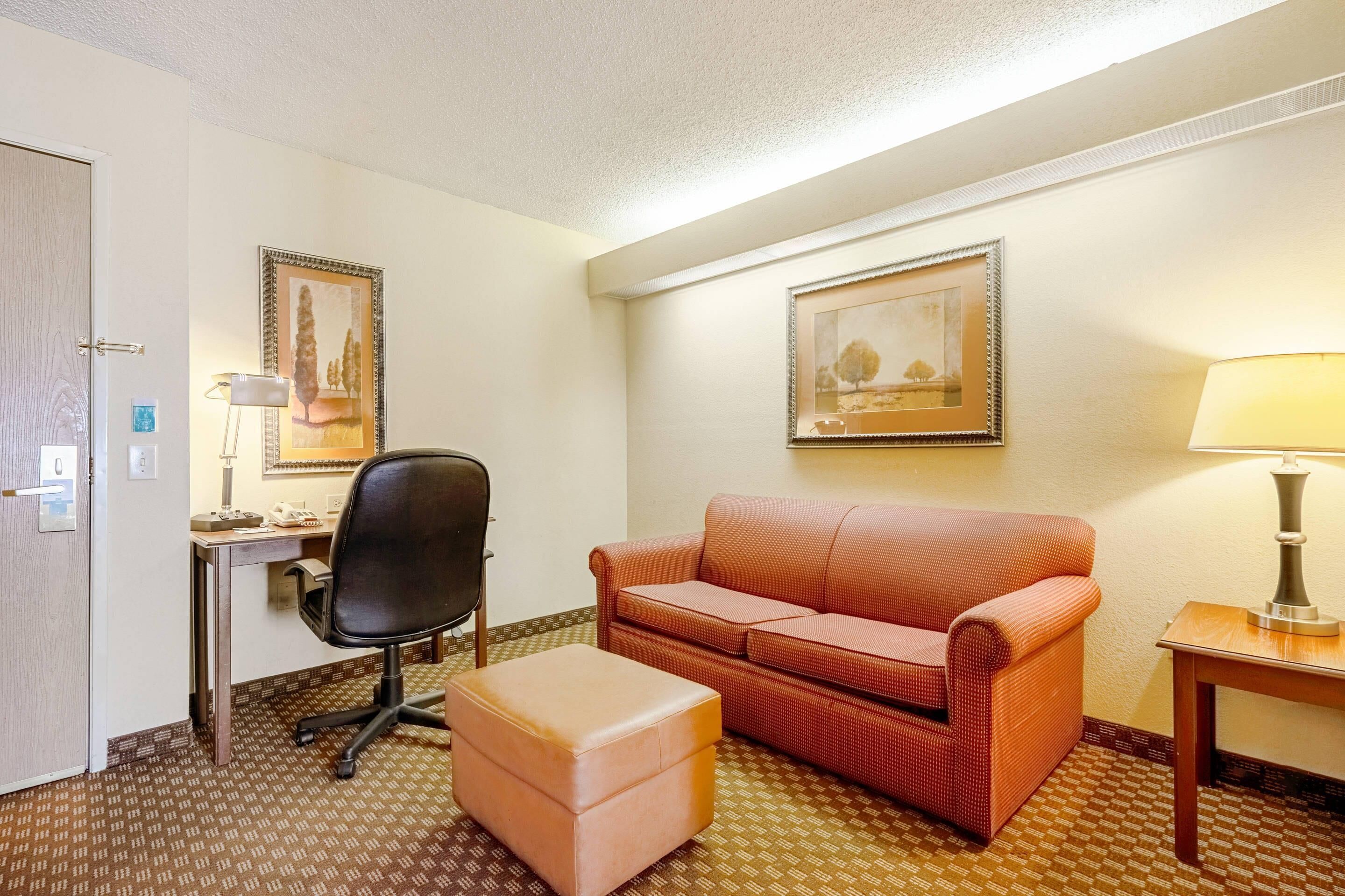 Habitación de hotel con escritorio, una silla, sofá, reposapiés, lámpara y dos cuadros en las paredes.