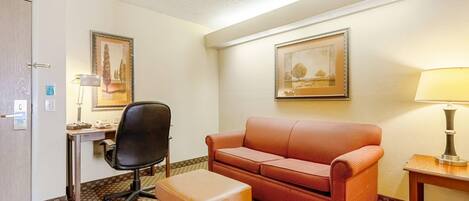 Habitación de hotel con escritorio, una silla, sofá, reposapiés, lámpara y dos cuadros en las paredes.