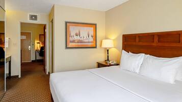 A hotel room with a large bed, a nightstand with a lamp, a framed picture on the wall, and a hallway leading to a bathroom.
