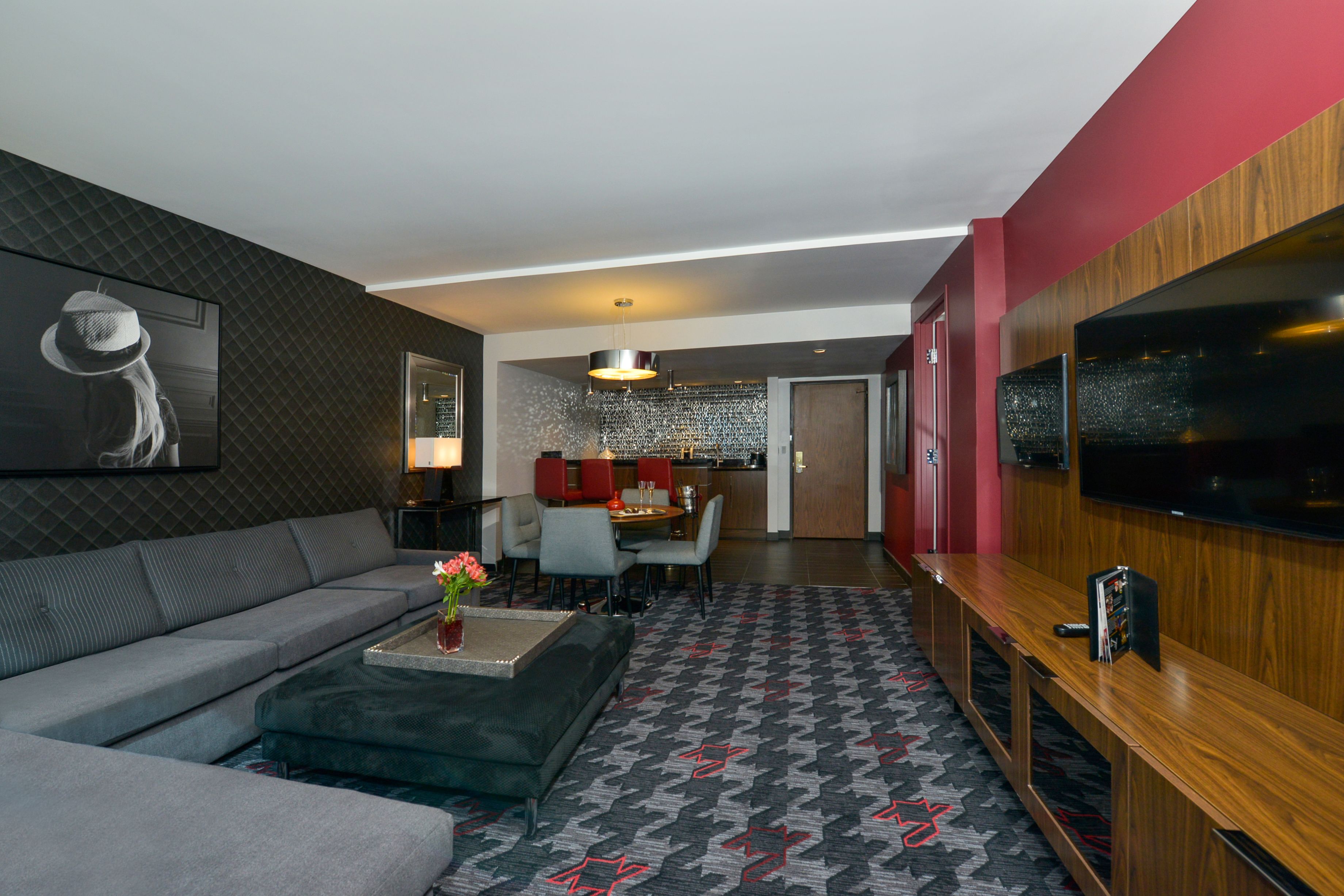 Una sala moderna con un sofá gris, una mesa de centro con flores, un tapete estampado, un mueble de televisión de madera y una zona de comedor al fondo.