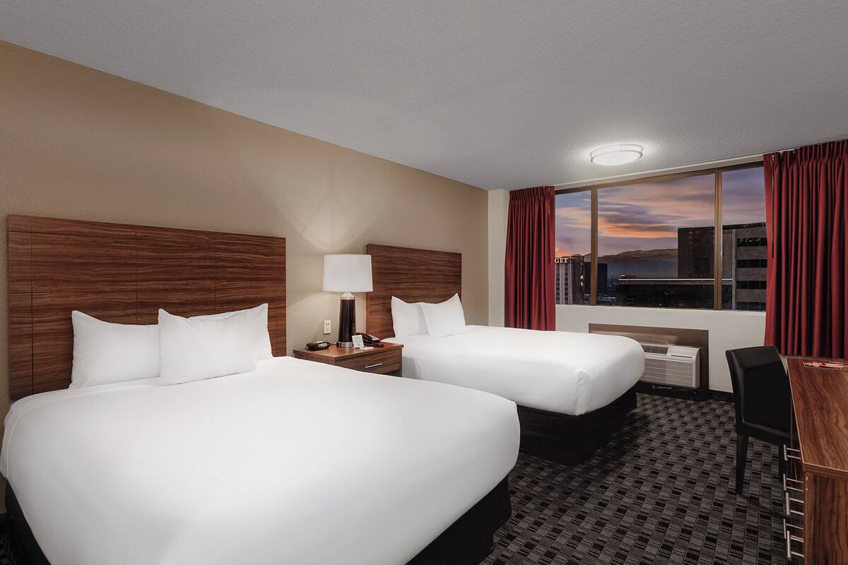 A hotel room with two beds, a desk, a chair, a lamp, and a view of the cityscape at sunset.