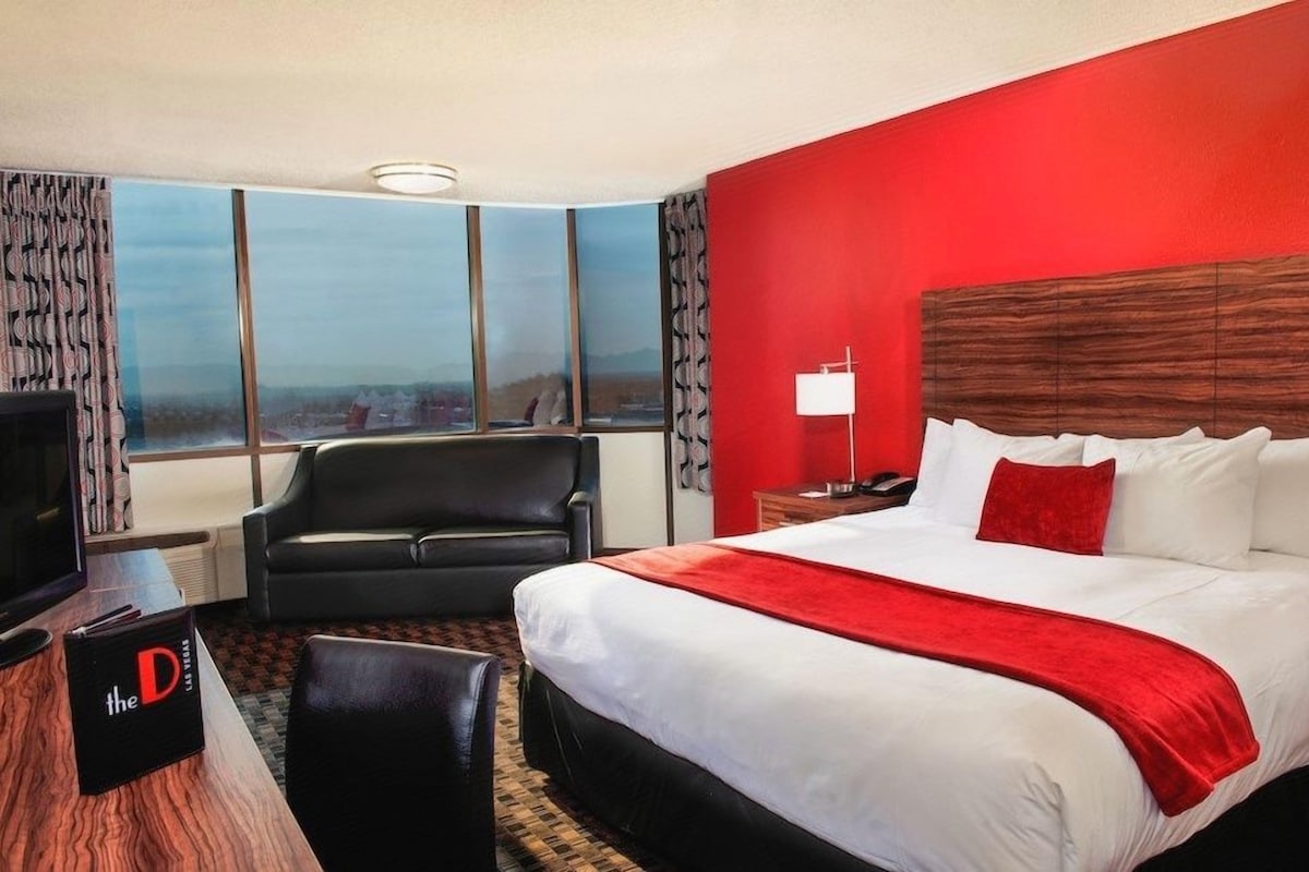 A hotel room with a large bed, a desk with a computer, a chair, and a view of the city through large windows.
