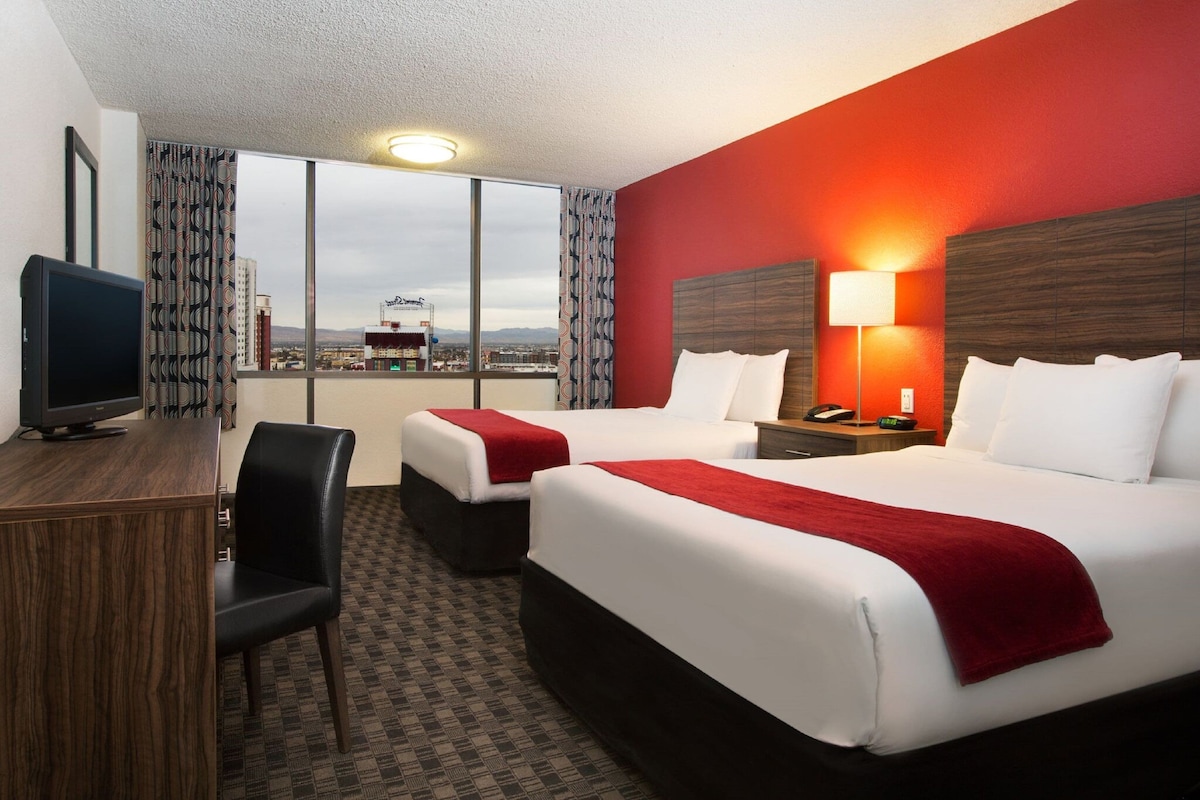 A hotel room with two beds, a desk, a chair, a TV, a lamp, and a city view.