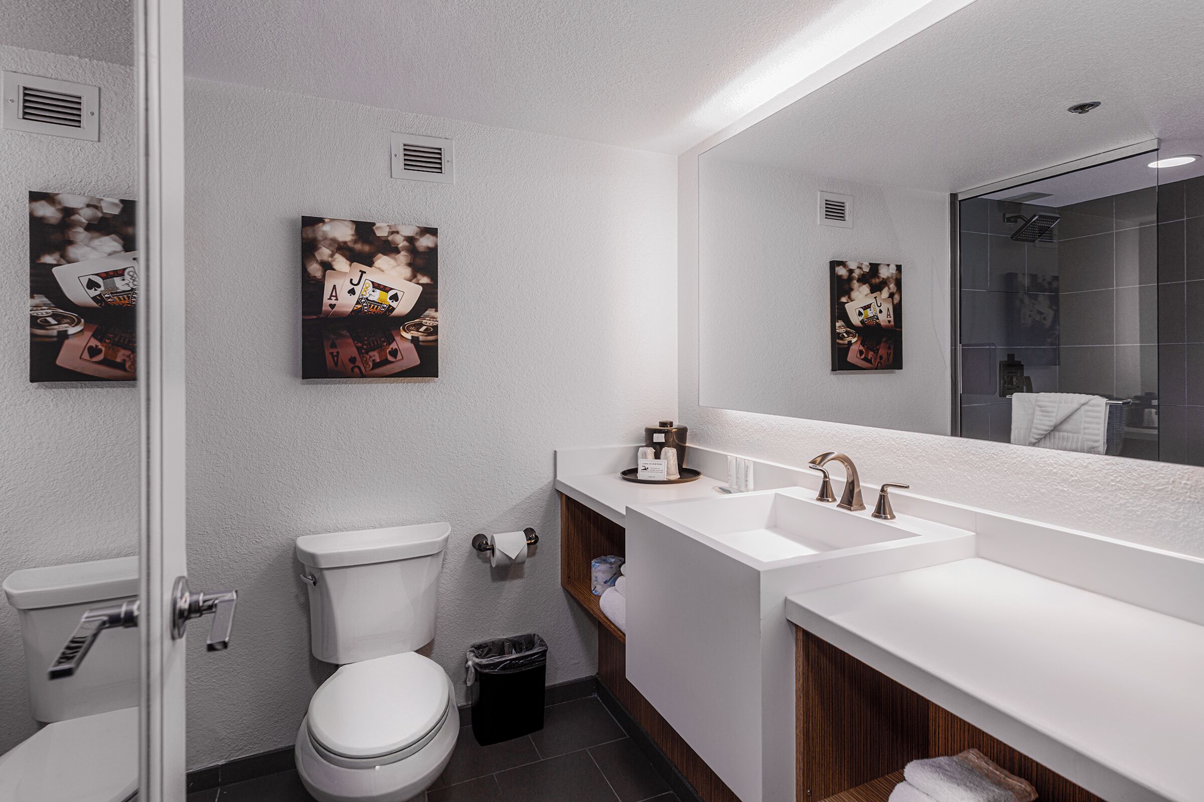 A modern bathroom with a white sink, dual faucets, and a large mirror. There are two framed artworks on the wall, a toilet, and a trash bin.