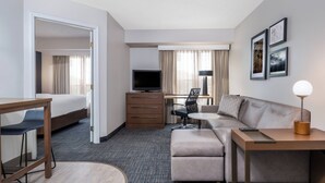 Ein Hotelzimmer mit einem Bett, einem Schreibtisch mit Stuhl, einer Couch, einem Fernseher, einer Lampe und gerahmten Bildern an der Wand.