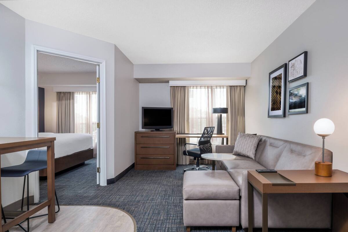 A hotel room with a bed, a desk with a chair, a sofa, a TV, a lamp, and framed pictures on the wall.