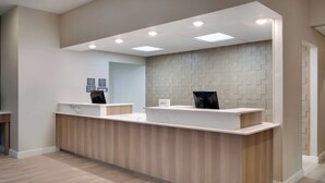 Ein Empfangsschalter mit heller Holzoptik, eine Wand mit geometrischem Muster aus Fliesen und ein Flachbildschirm.