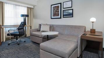Habitación de hotel con sofá, una pequeña mesa redonda, escritorio con lámpara, una silla y ventana con cortinas.