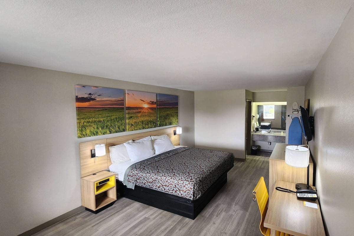 A hotel room with a bed, bedside tables, a desk with a chair, a mirror, and a painting of a sunset over a field.
