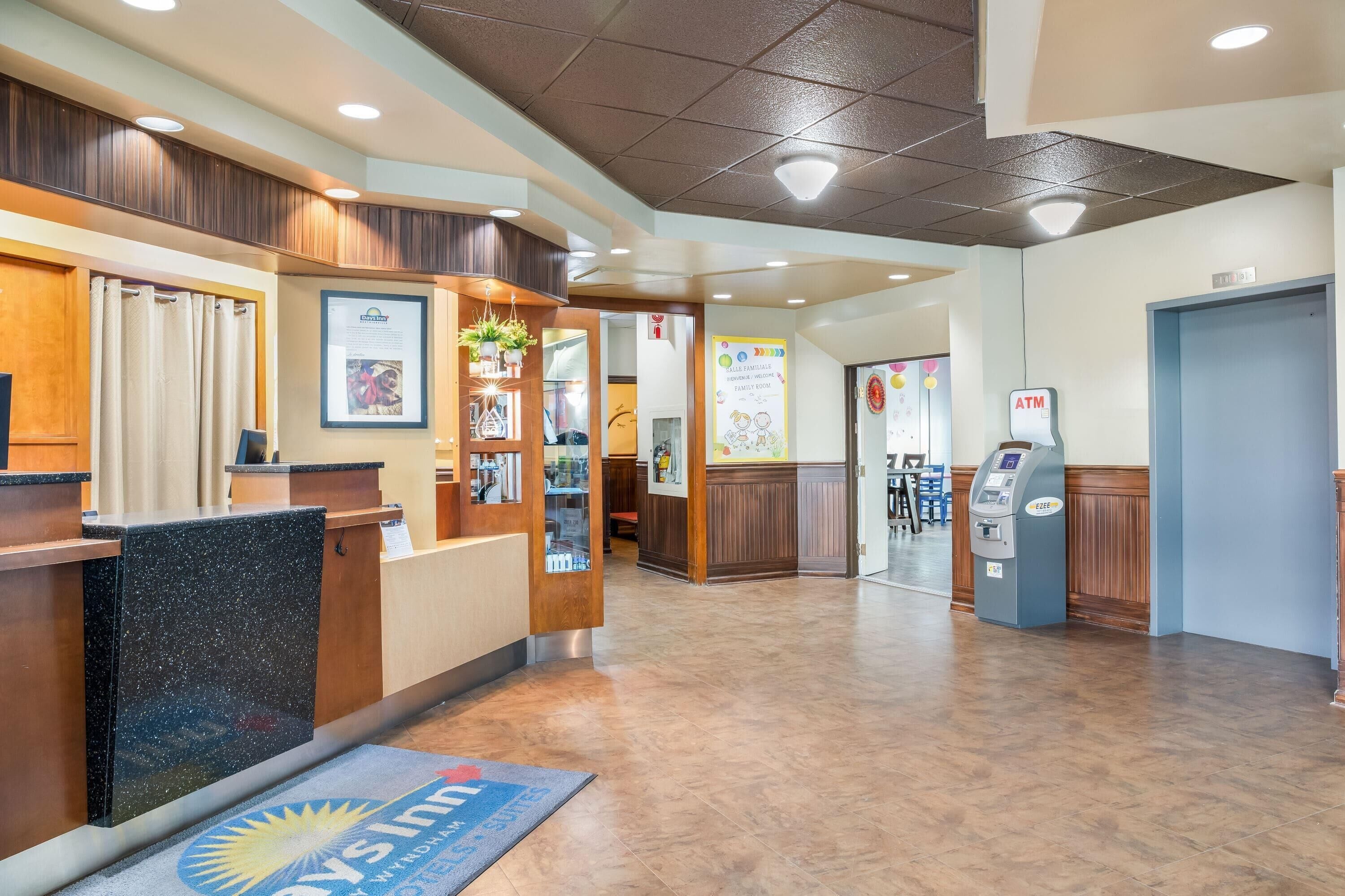 A hotel reception area with a reception desk, a blue mat with the hotel's logo, an ATM, and a bulletin board with various notices.