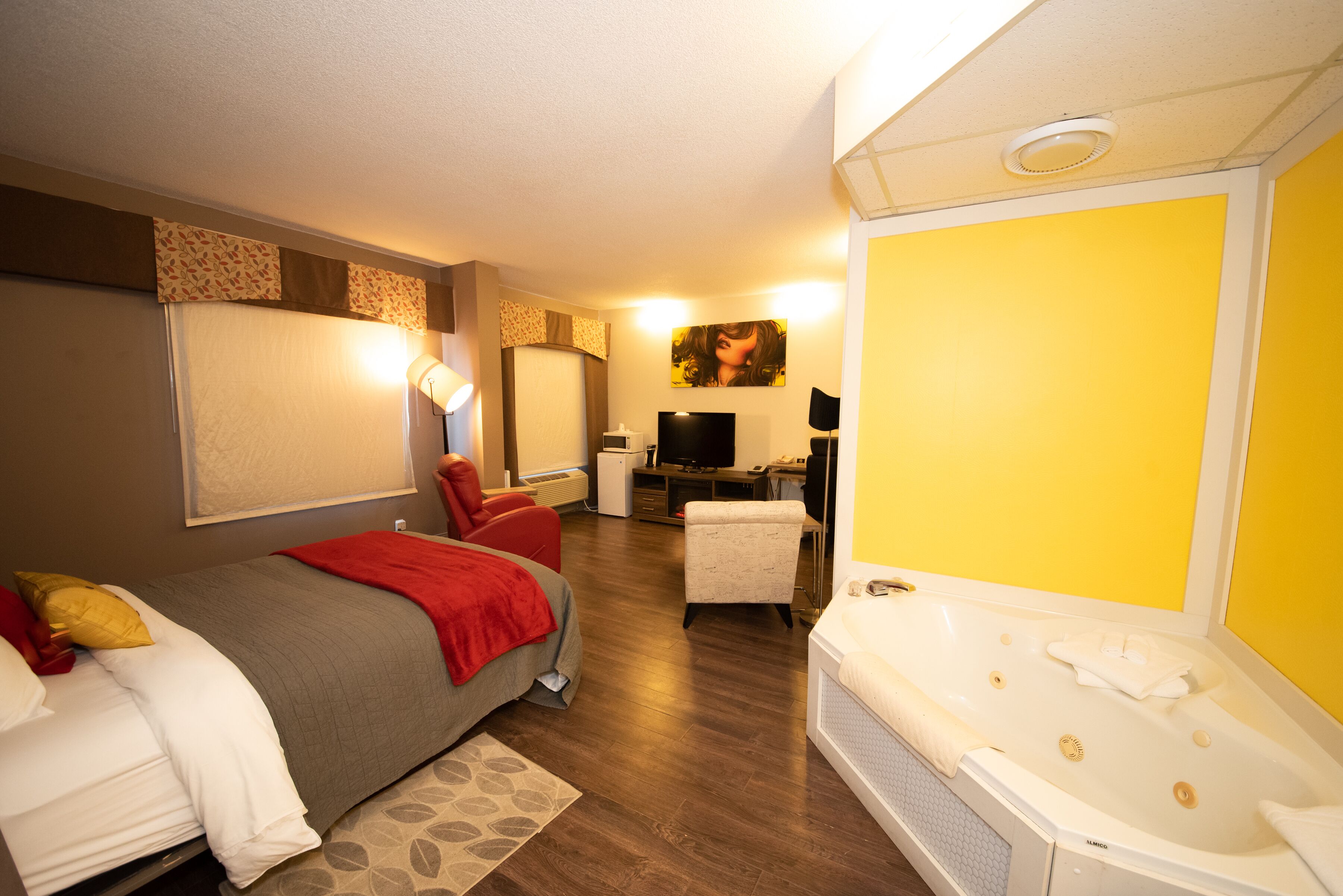 A hotel room with a bed, a red chair, a desk with a computer, a TV, and a large bathtub.