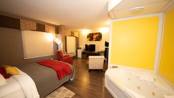 A hotel room with a bed, a red chair, a desk with a computer, a TV, and a large bathtub.