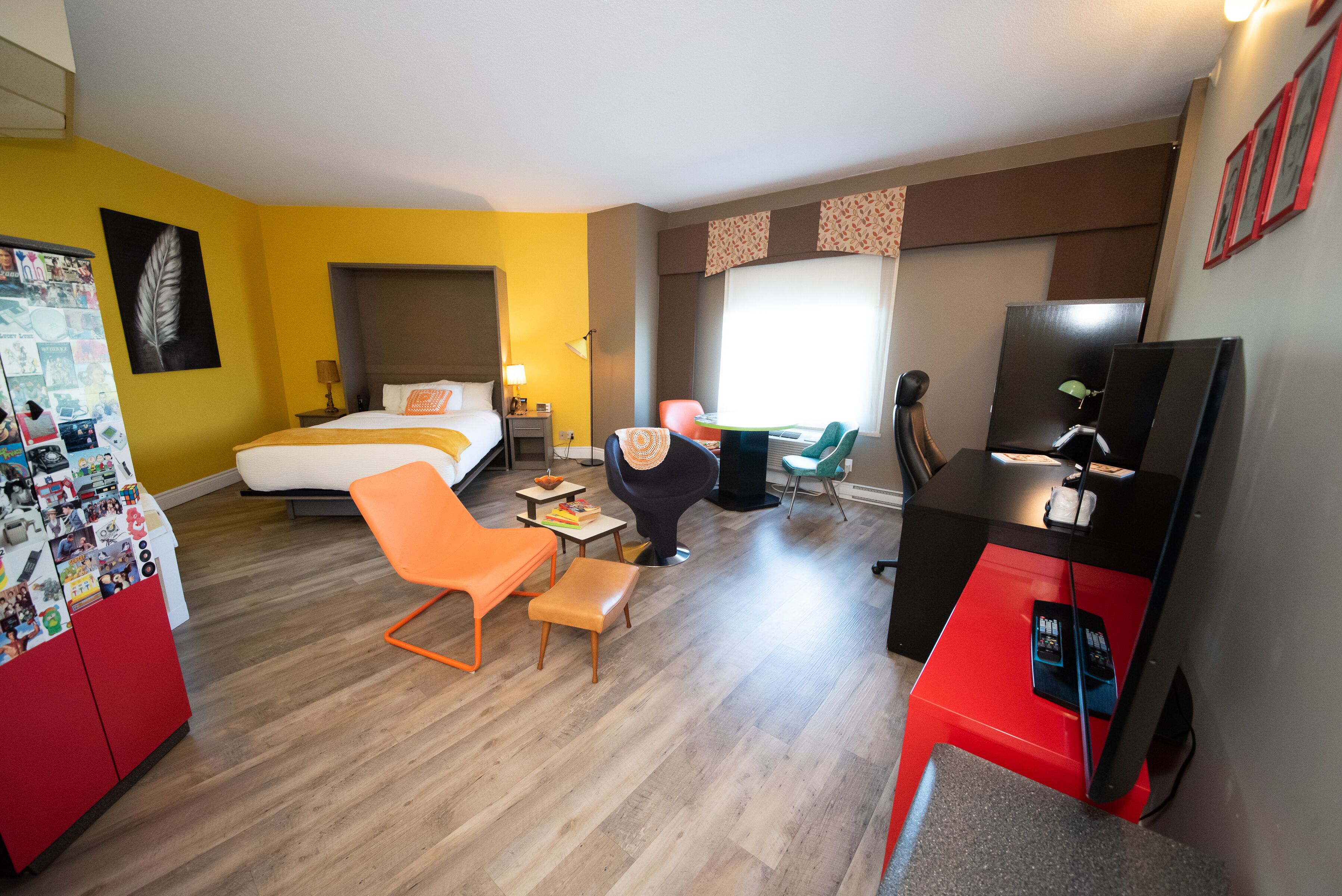 Quarto de hotel com cama, poltrona laranja, escrivaninha e televisão.