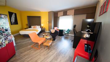 Quarto de hotel com cama, poltrona laranja, escrivaninha e televisão.