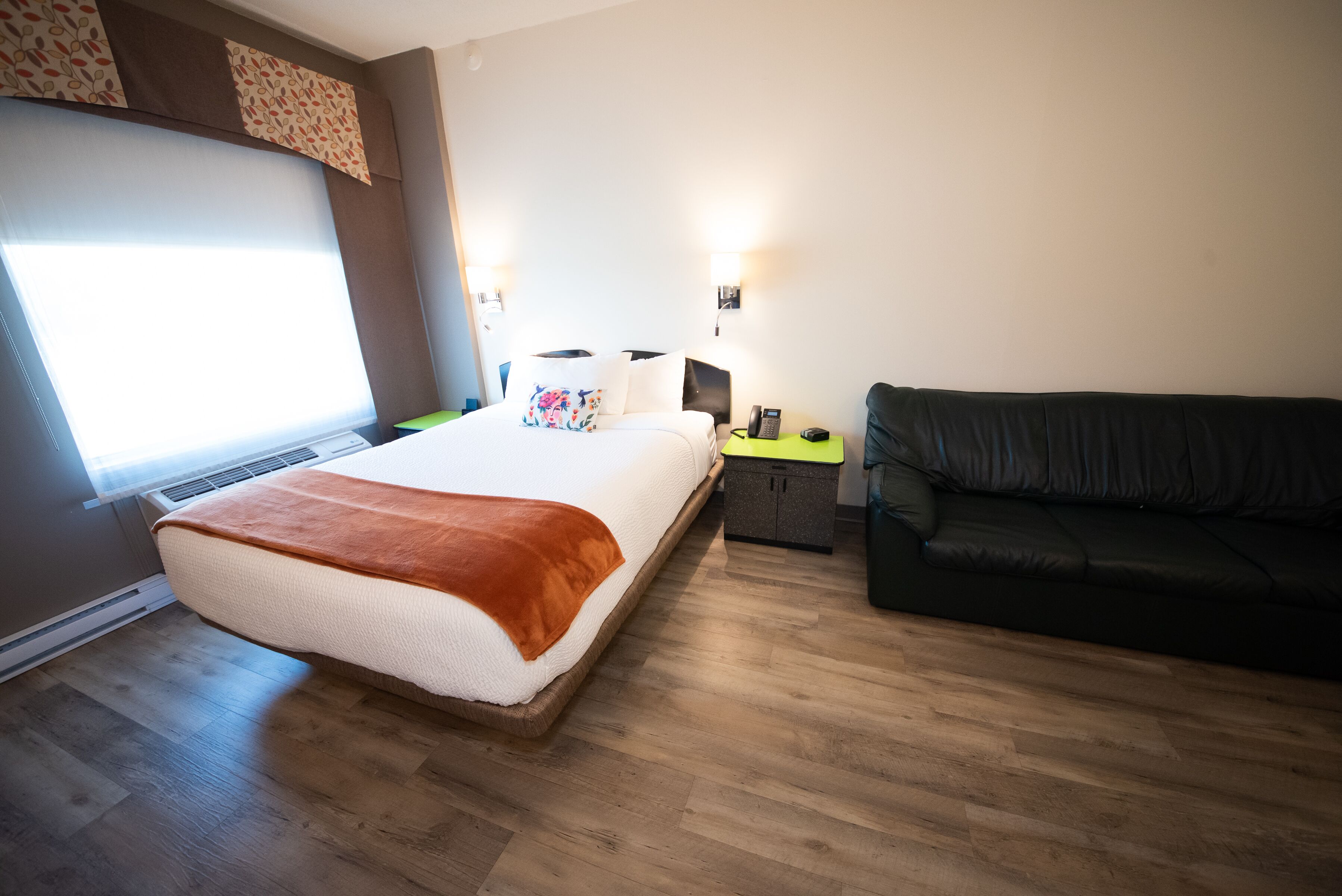 Une chambre d’hôtel avec un grand lit, un canapé en cuir noir, une table de chevet avec un téléphone et une fenêtre équipée de stores.