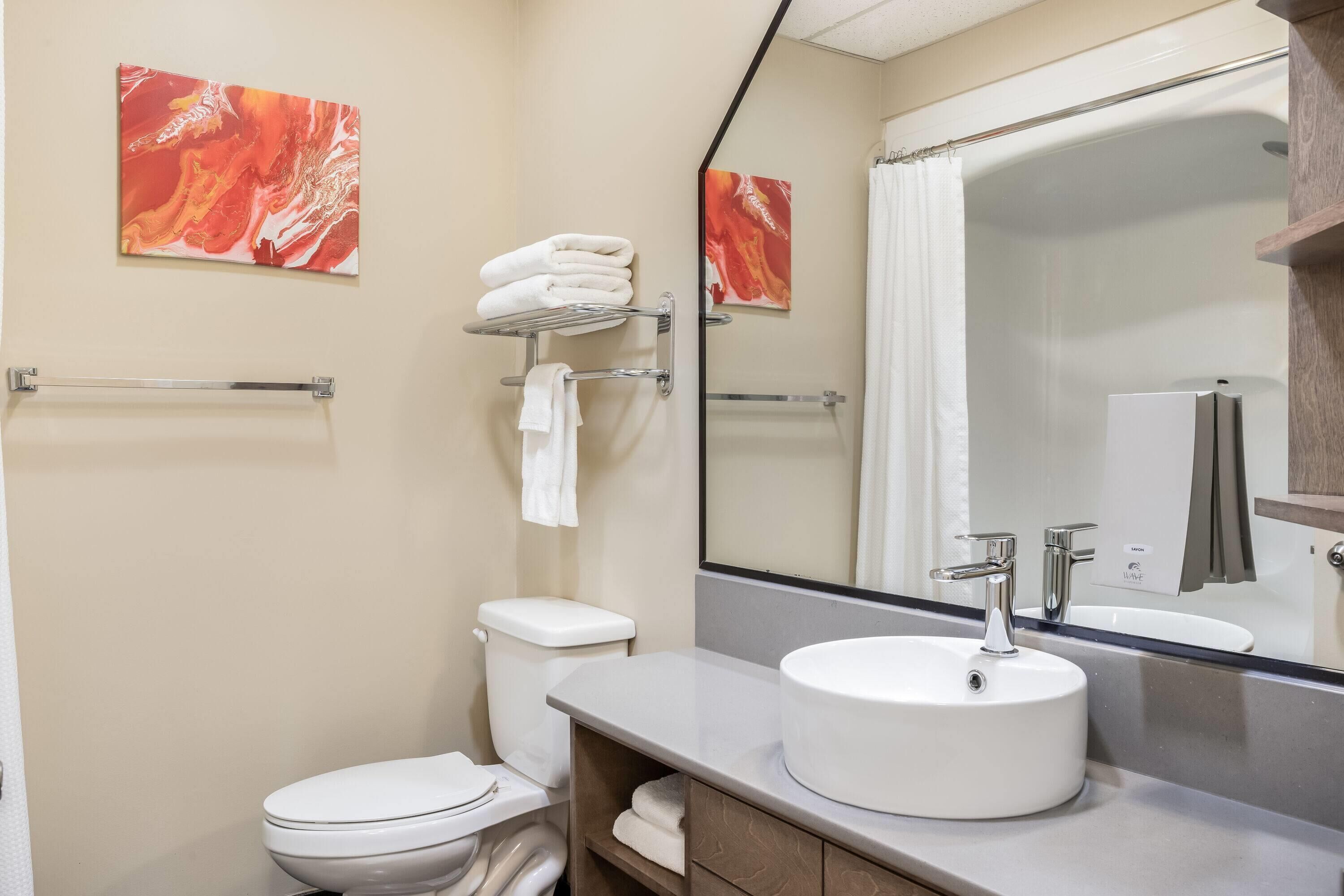 Un baño moderno con inodoro blanco, un lavabo con espejo superior y dos cuadros abstractos en la pared.