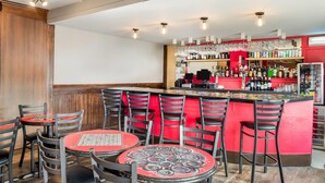 Zona de bar con varias mesas y sillas, barra con diferentes botellas e iluminación colgante.