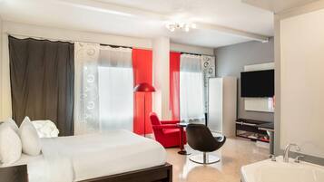 Habitación de hotel con cama, una silla roja, una silla negra, televisor y baño con lavamanos.