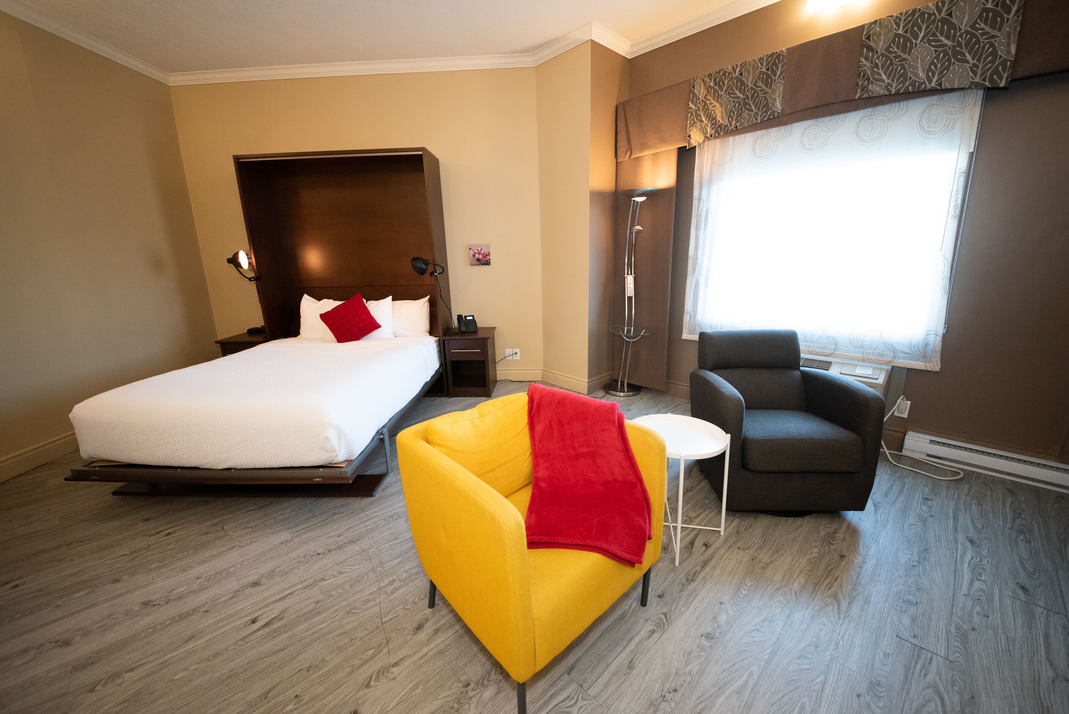A hotel room with a bed, a yellow armchair, a grey sofa, a small table, and a shower area.