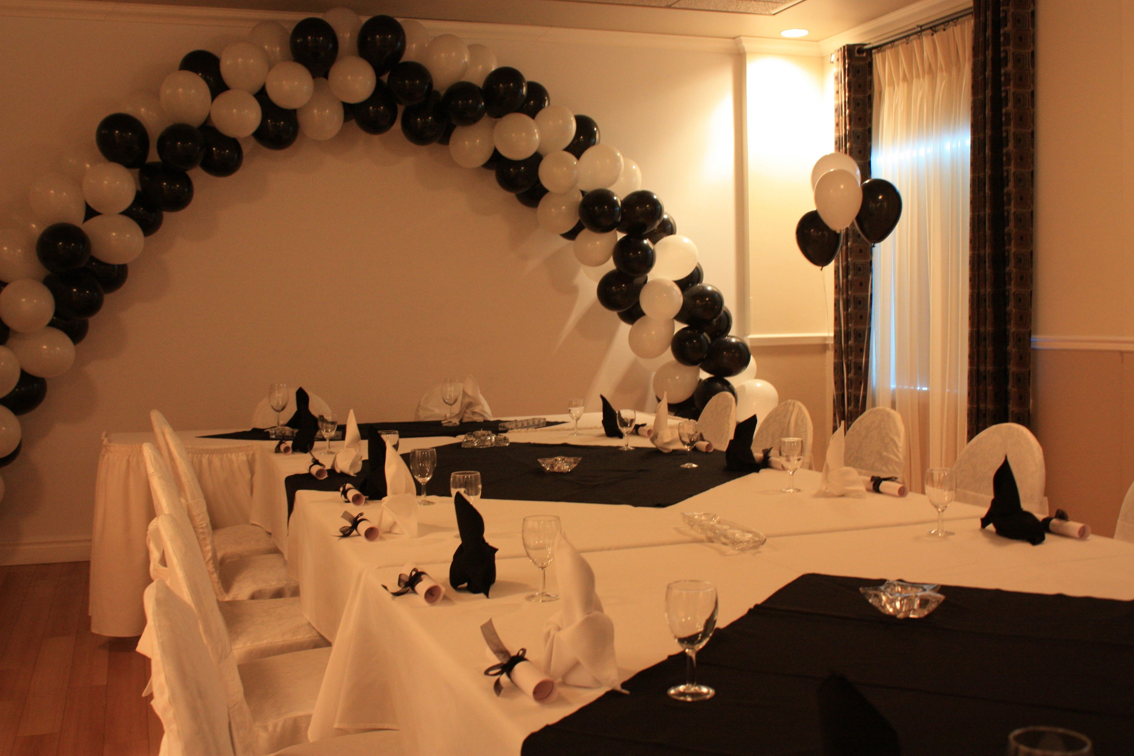 A dining area with tables set for a formal event, featuring white and black balloons forming an arch.