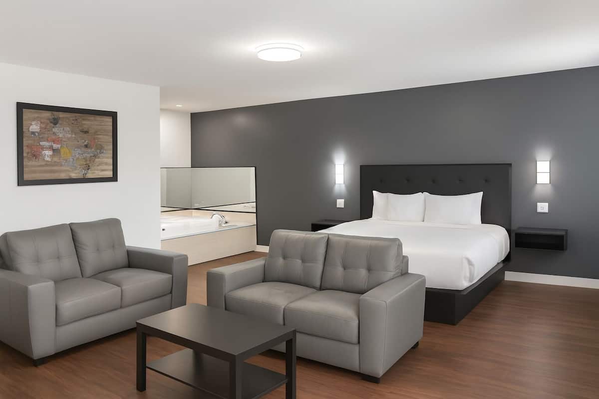 A modern hotel room with a large bed, two armchairs, a small table, a bathroom with a mirror, and a painting on the wall.