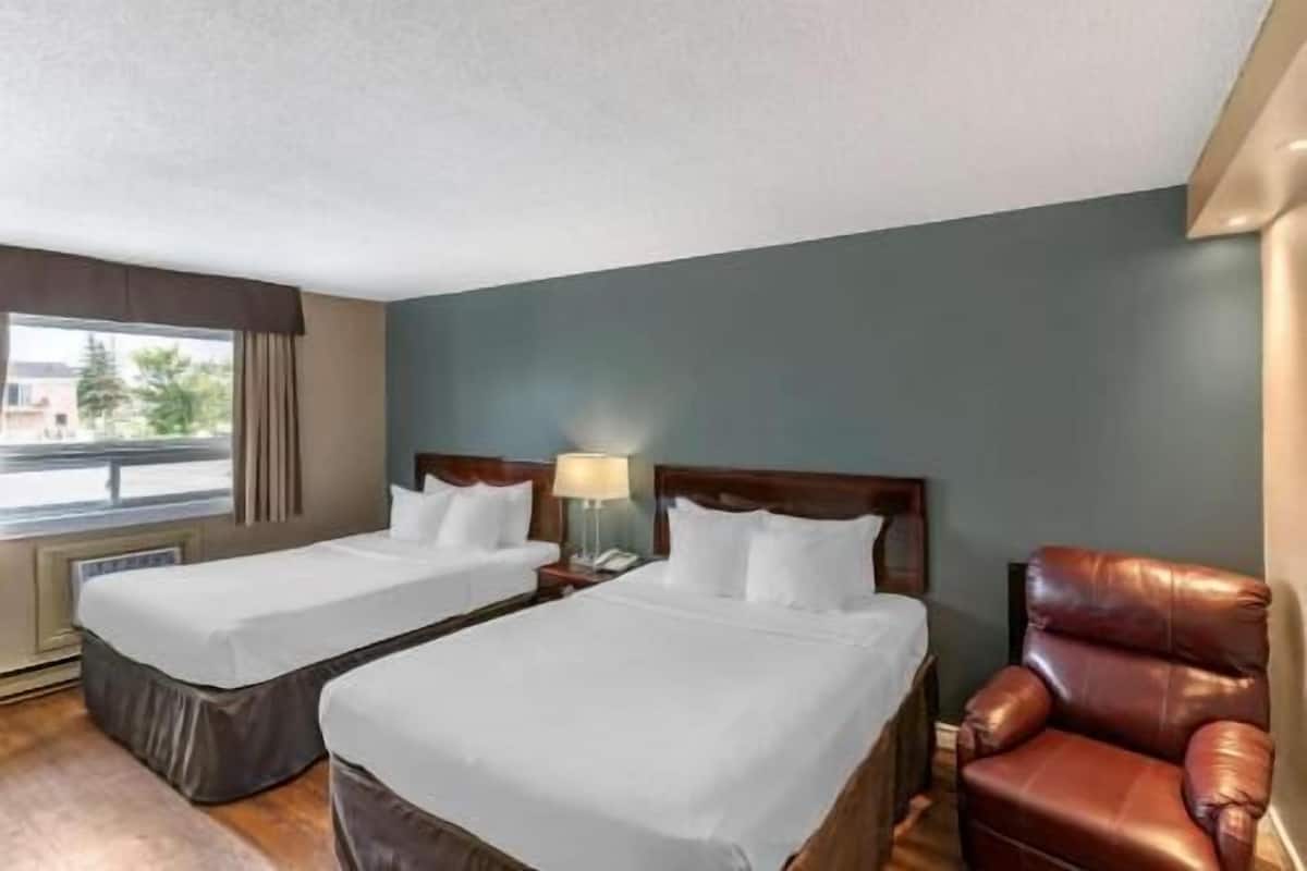 A hotel room with two beds, a chair, a lamp, and a window with a view.