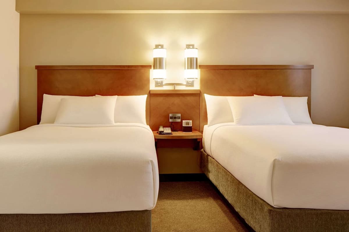 A hotel room with two beds, a wooden headboard, a nightstand with a phone and a lamp, and a carpeted floor.