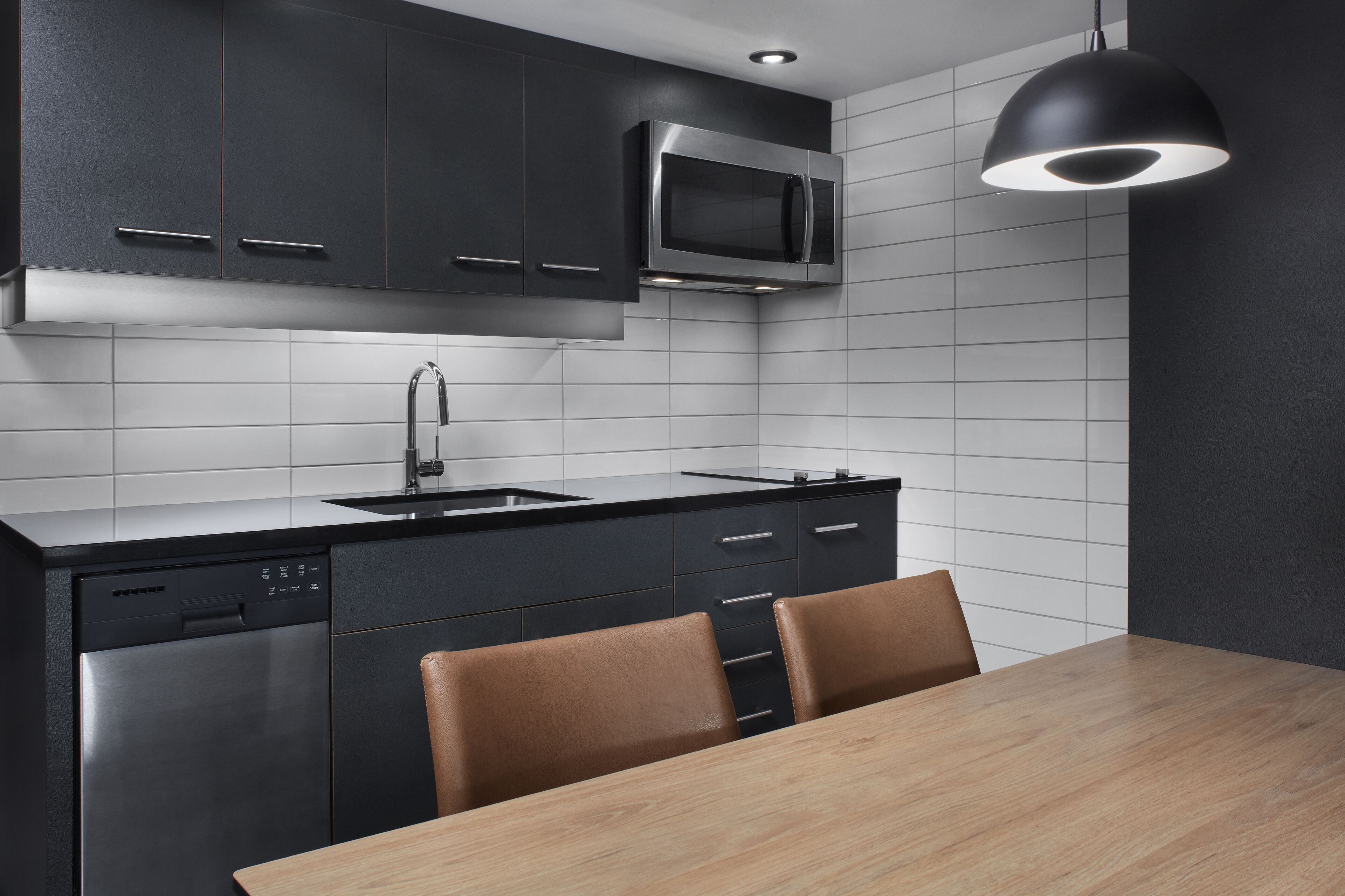 A modern kitchen with dark cabinetry, a stainless steel dishwasher, a microwave, and a wooden dining table with chairs.
