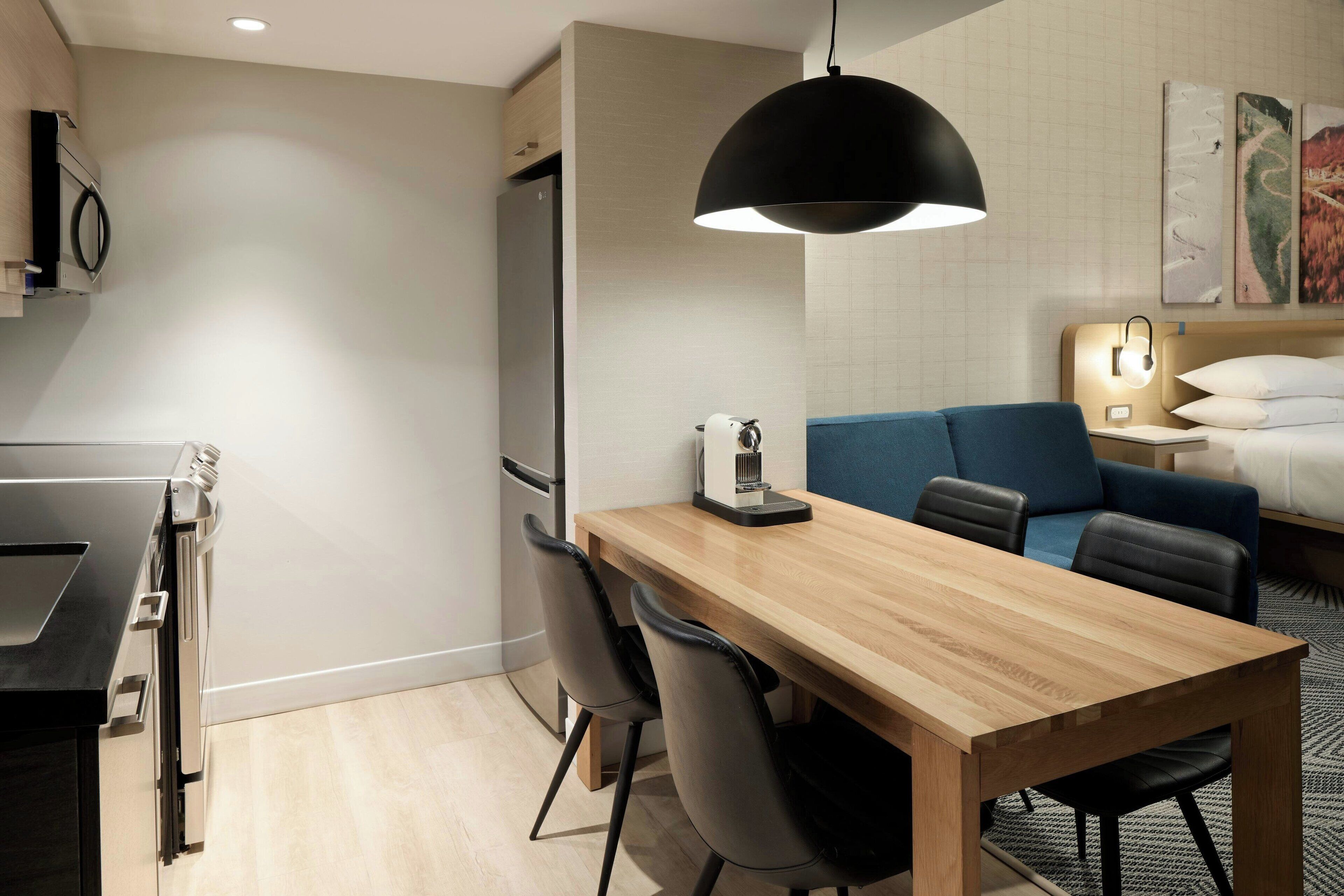 A modern hotel room with a wooden dining table, black chairs, a blue sofa, and a bed with white linens.