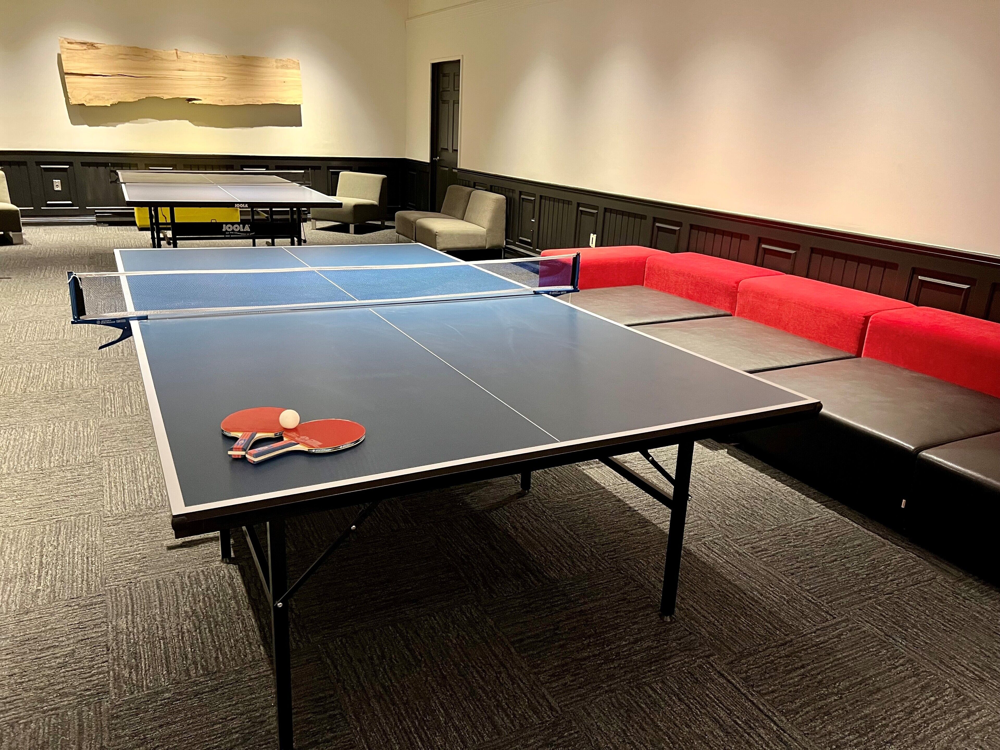 Una mesa de ping pong con dos paletas y una pelota, un sofá rojo y un adorno de pared de madera en una habitación con suelo alfombrado.