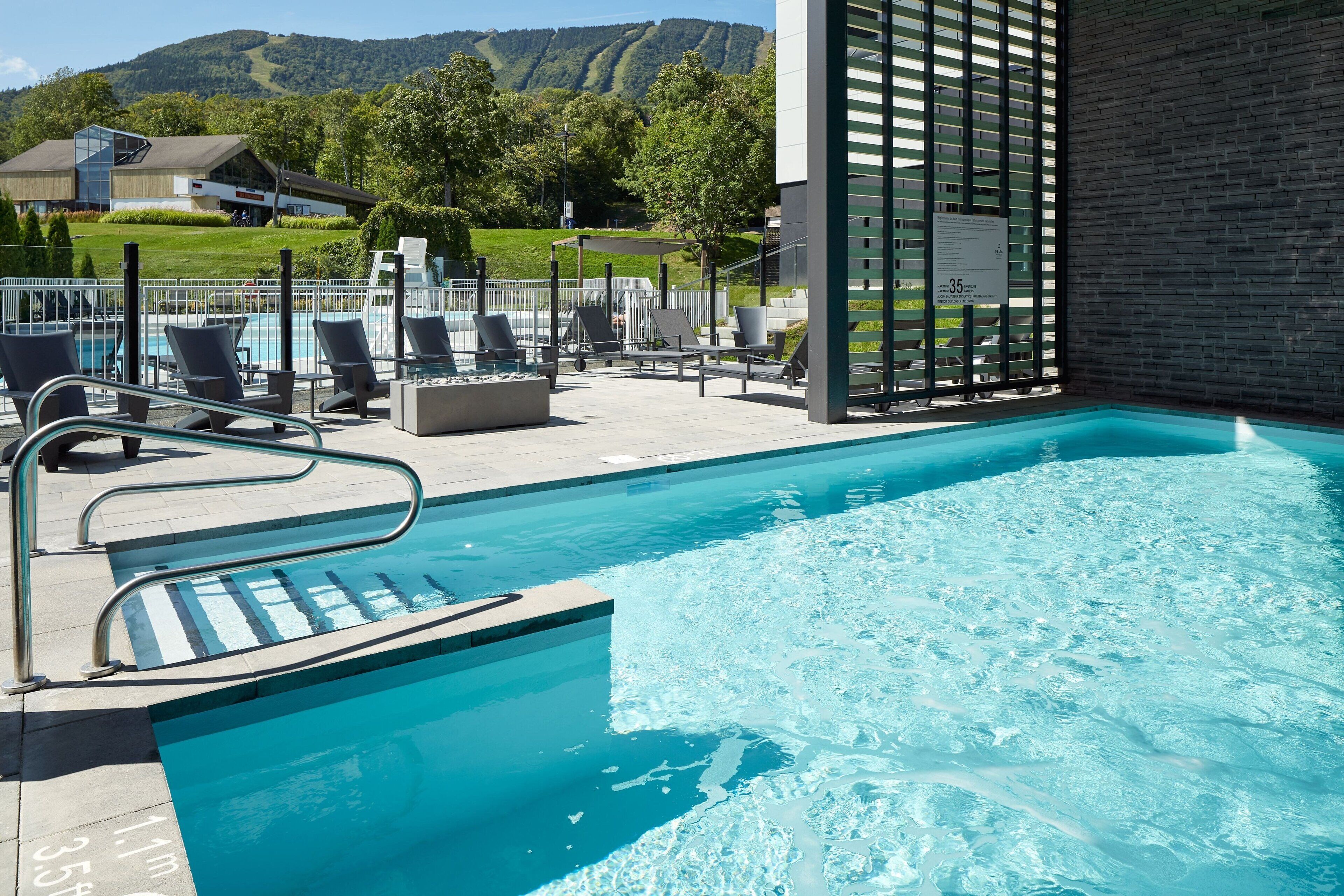 Une piscine avec des chaises longues, donnant sur un bâtiment et des collines en arrière-plan.