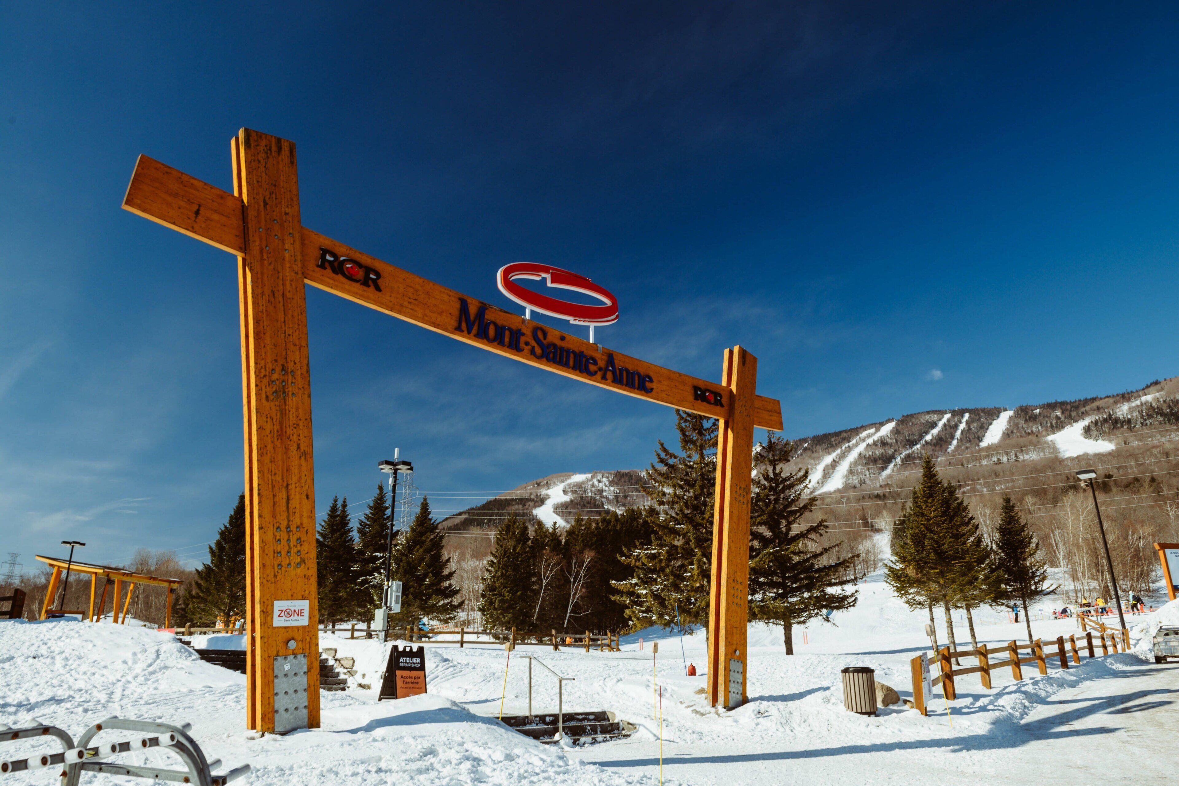Ein hölzernes Schild mit der Aufschrift 'Mont-Sainte-Anne' und 'RCR' vor einer verschneiten Landschaft mit einem Berg im Hintergrund.