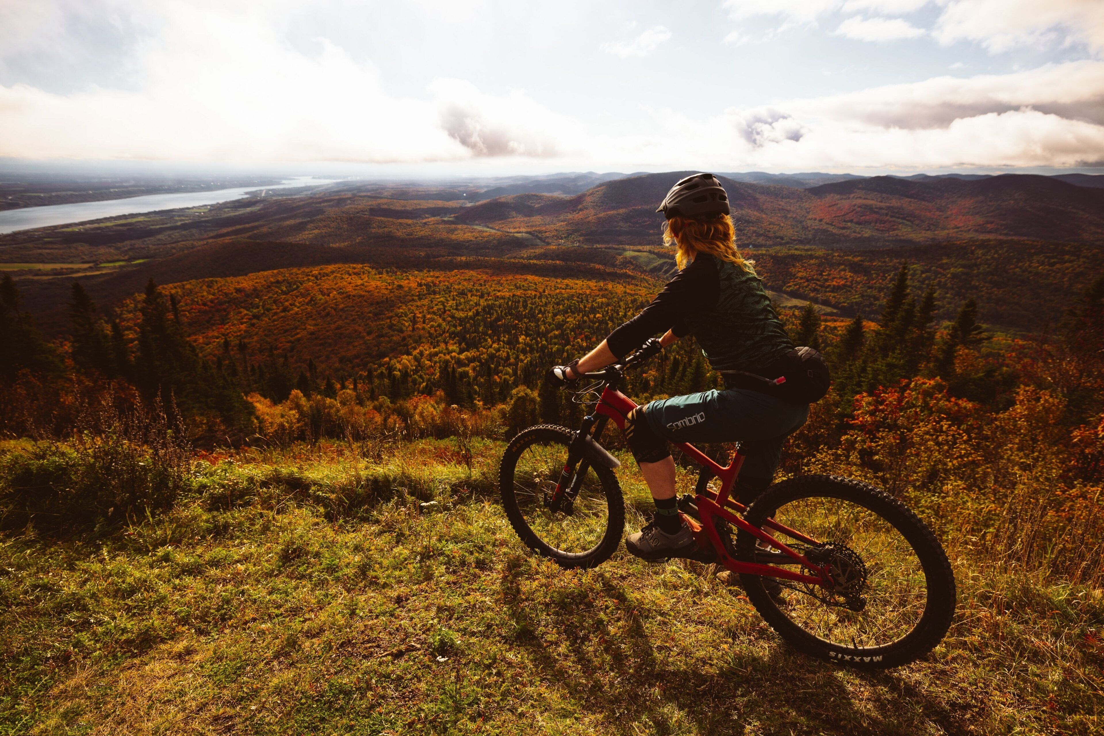 Una persona pedalea en bicicleta de montaña por una colina cubierta de hierba, con una vista panorámica de un valle y montañas al fondo.