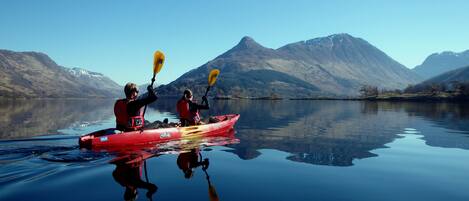 Dos personas en kayak en un lago tranquilo, con montañas al fondo.