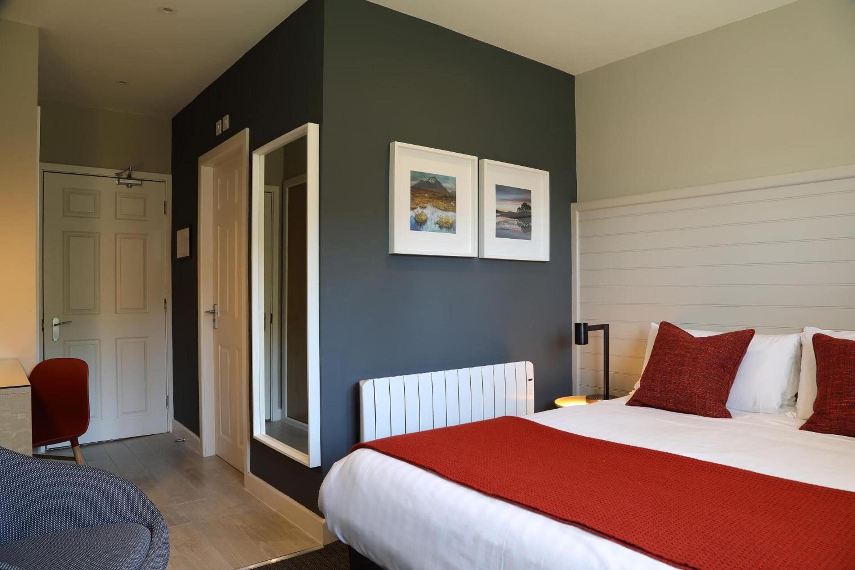 A hotel room with a bed, a red chair, a white radiator, and two framed pictures on the wall.