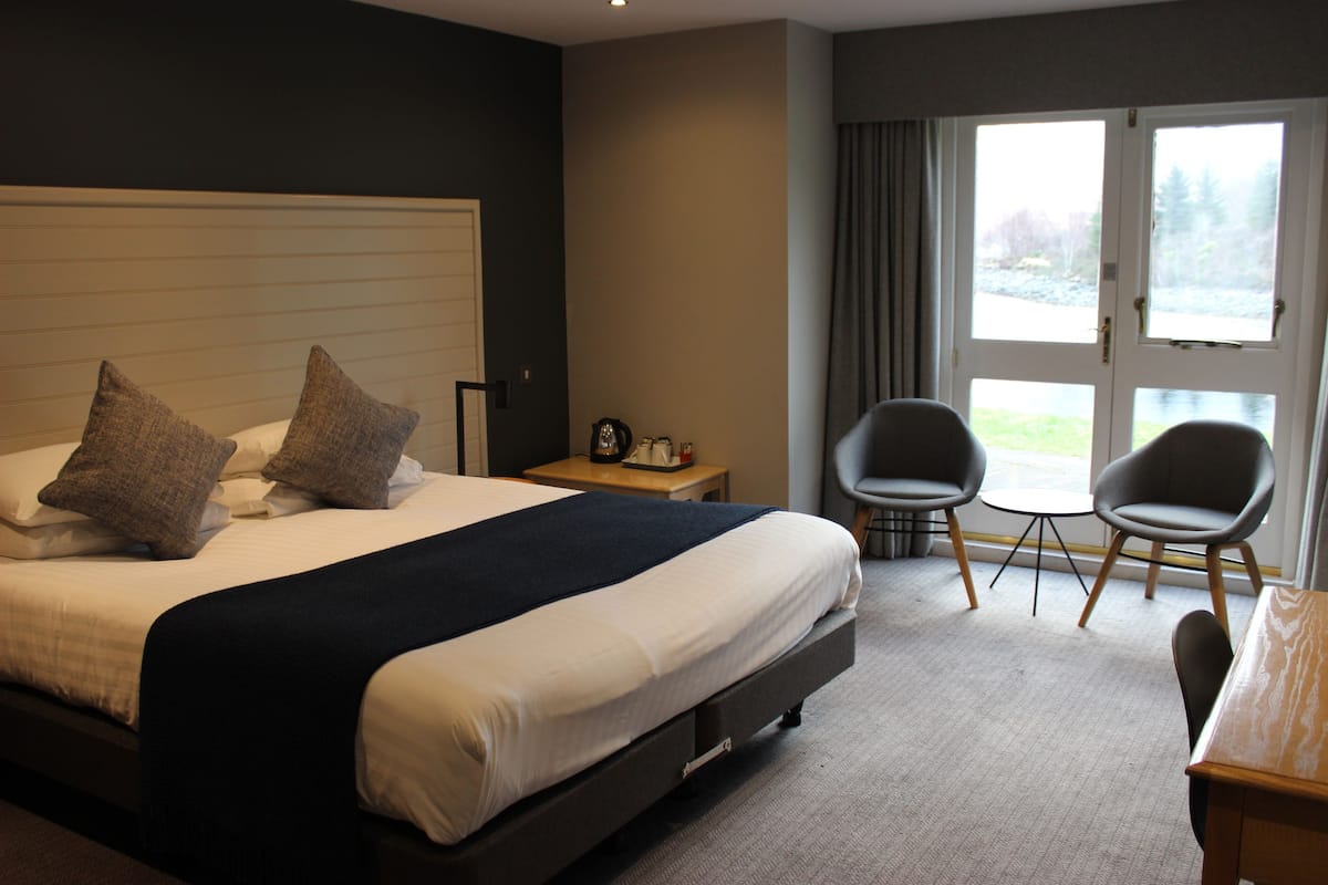 A hotel room with a large bed, a small table, two chairs, and a view of the outdoors through a door.