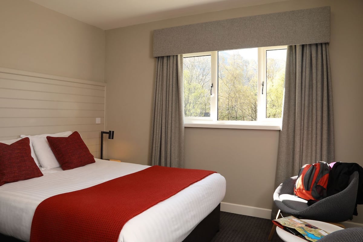 A hotel room with a bed, a window with curtains, a chair, and a table with a book.