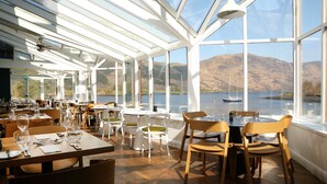 Restaurante com grandes janelas que oferecem vista para um lago e montanhas. Mesas com cadeiras e talheres.
