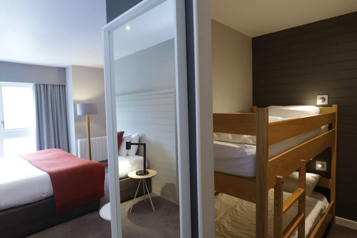 A hotel room with a bunk bed, a single bed, a small table with a lamp, and a large mirror.