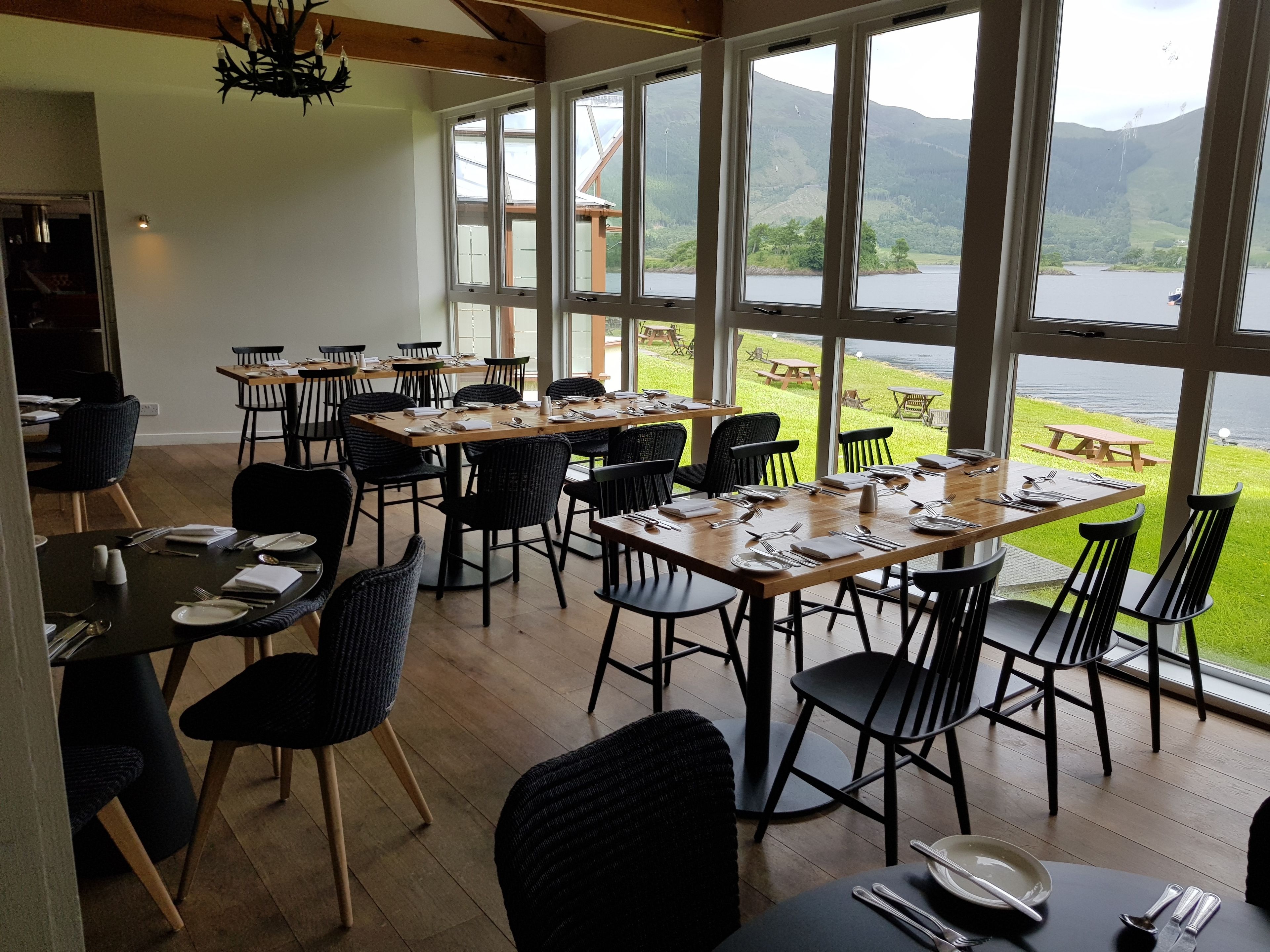 A dining area with tables set for four, large windows offering a view of a lake and mountains.