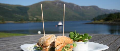 Un sandwich sur une assiette blanche, accompagné d’un petit bol de salade verte, le tout posé sur une table en bois, avec un lac et des montagnes en arrière-plan.