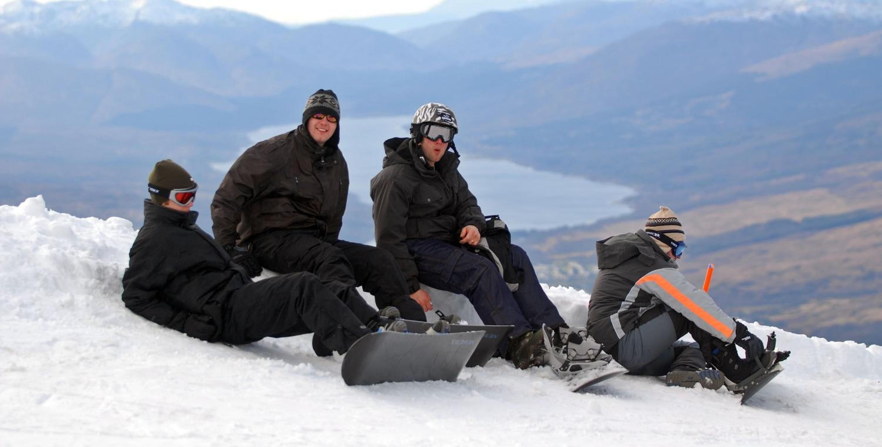 Four people sitting on a snowy mountain with snowboards, surrounded by a mountainous landscape.