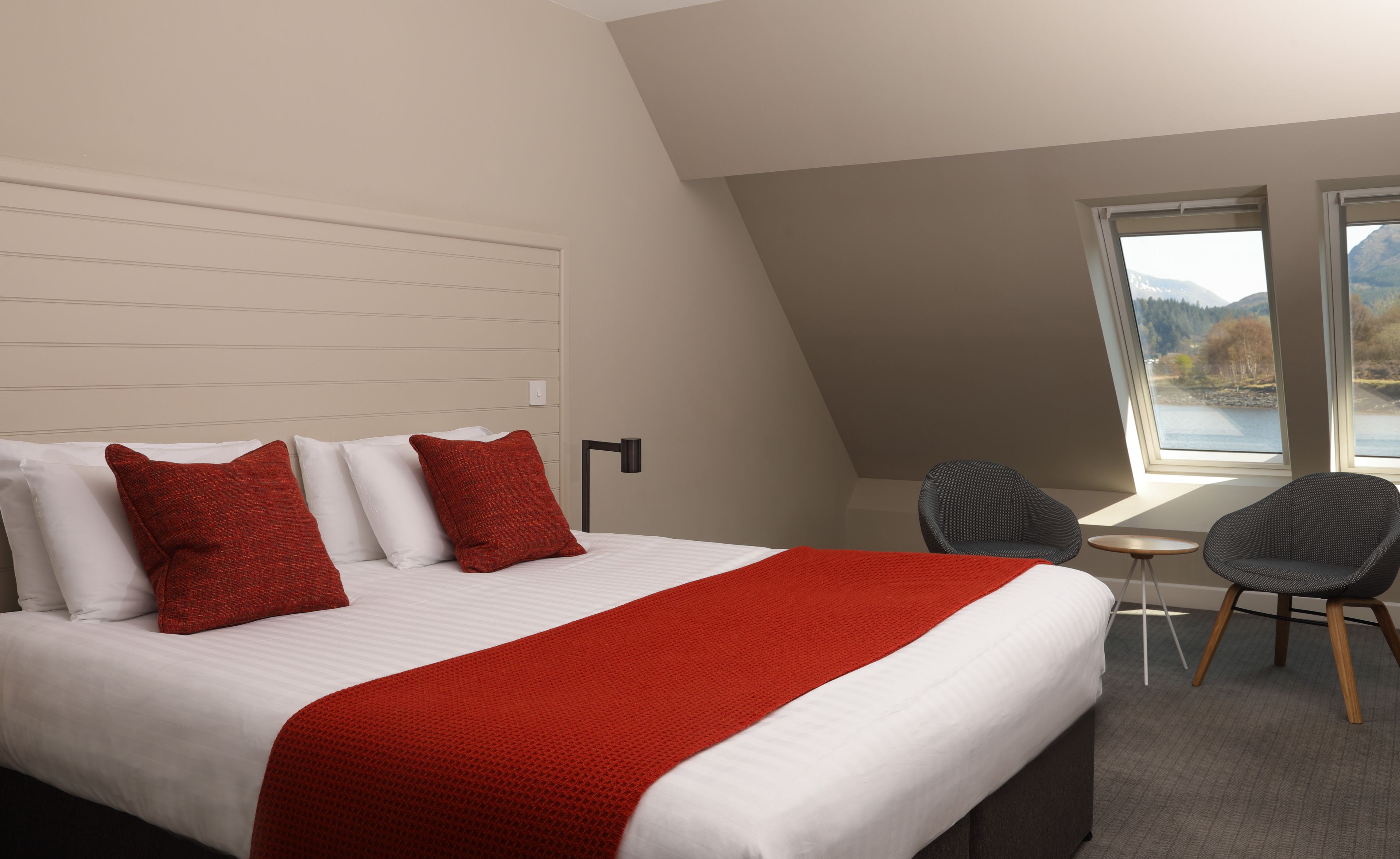 Una habitación de hotel moderna con una cama grande, ropa de cama blanca y roja, una pequeña mesa, dos sillas y vistas a un lago a través de un ventanal amplio.