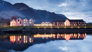 A large, multi-story building with a symmetrical facade, reflecting in a calm body of water, surrounded by mountains. - Isles Of Glencoe Hotel (Ballachulish)