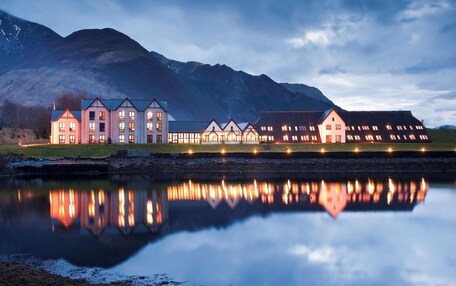 A large, multi-story building with a symmetrical facade, reflecting in a calm body of water, surrounded by mountains.