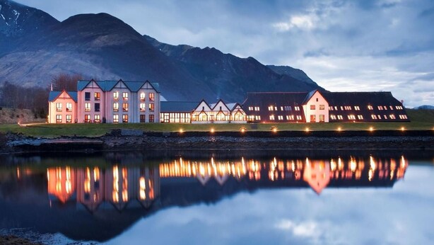 Um edifício grande e de vários andares com fachada simétrica, refletido em águas calmas, cercado por montanhas.