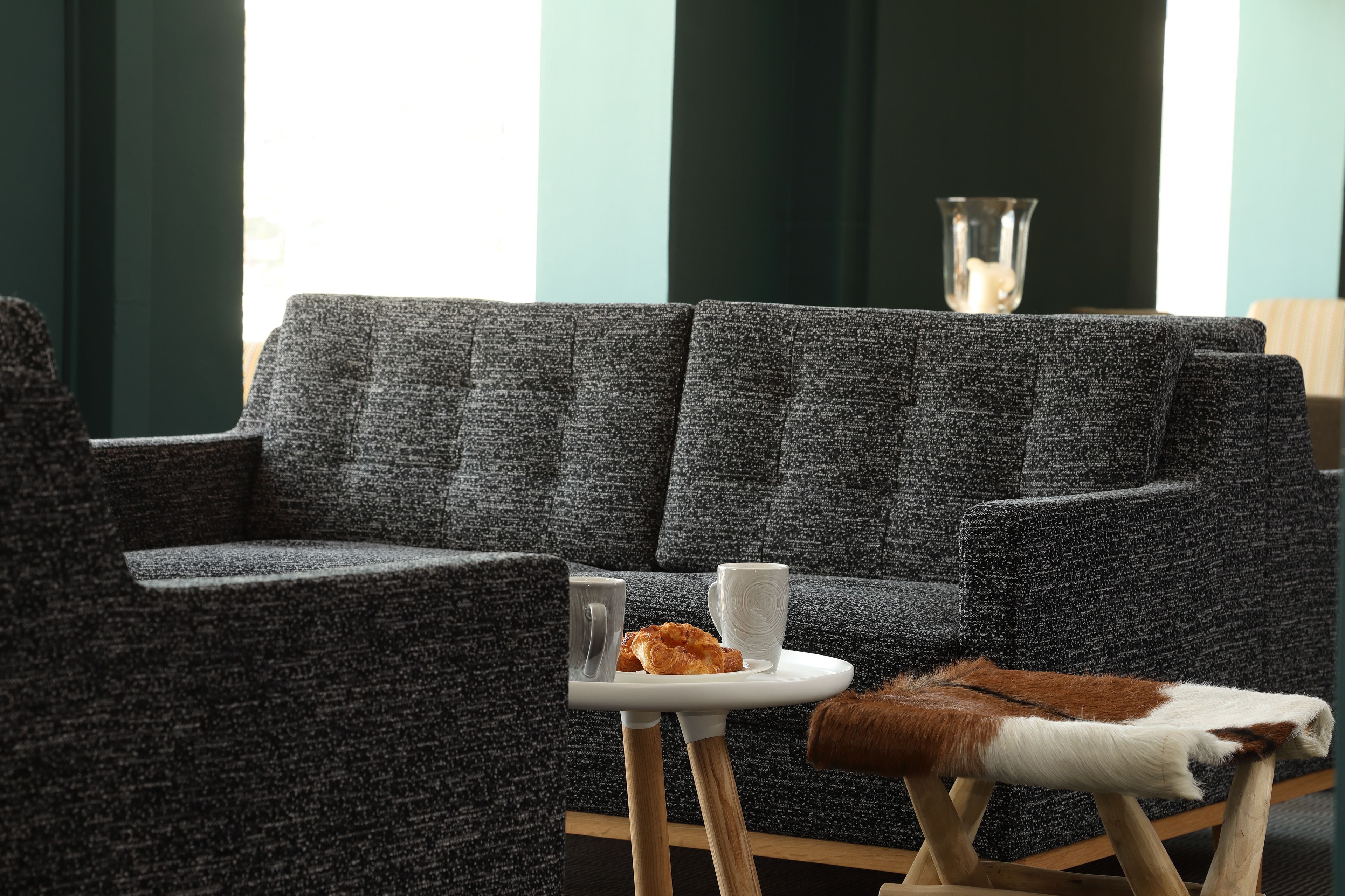 Una sala de estar moderna con un sofá gris, una mesita blanca con una taza y un pastel, y un jarrón de vidrio sobre un pedestal.