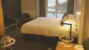 Quarto de hotel com cama, escrivaninha com telefone e luminária, cadeira, mesa pequena com chávenas e vista para o mar através de amplas janelas.