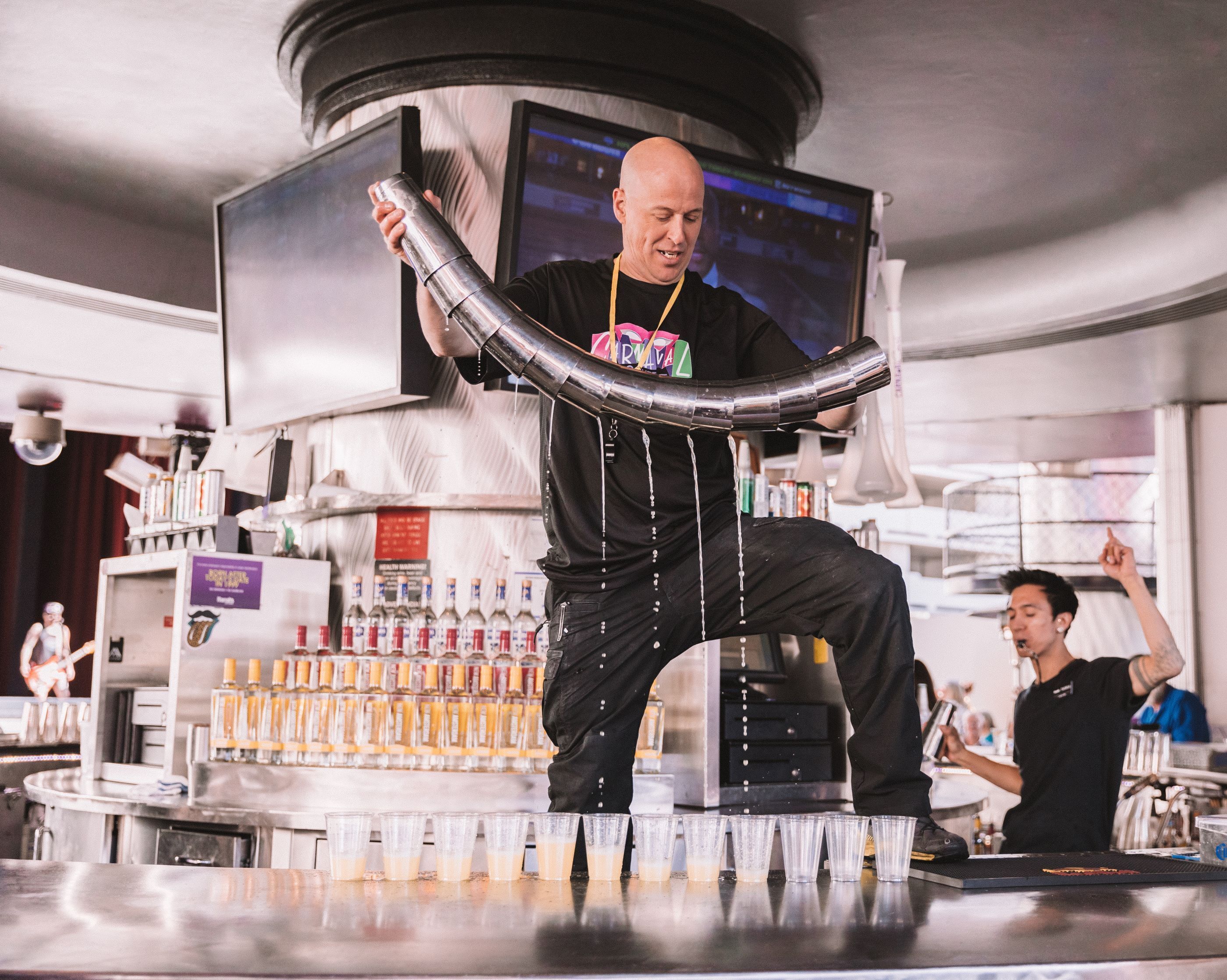 A bar area with a bartender preparing drinks, a counter with bottles, and a television screen in the background.